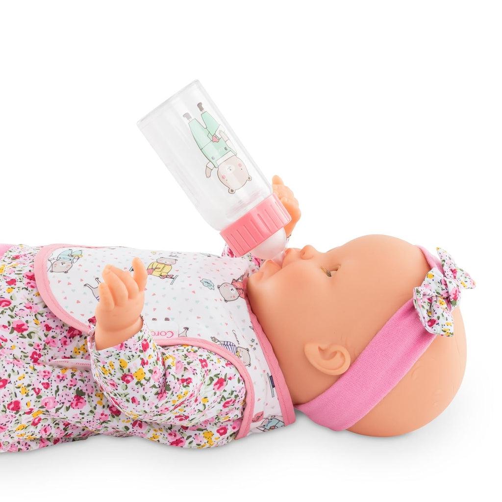 A baby doll is shown on it's back with the bib on and the bottle in it's mouth, the "milk" in the bottle appears to be empty due to the nature of the bottle.