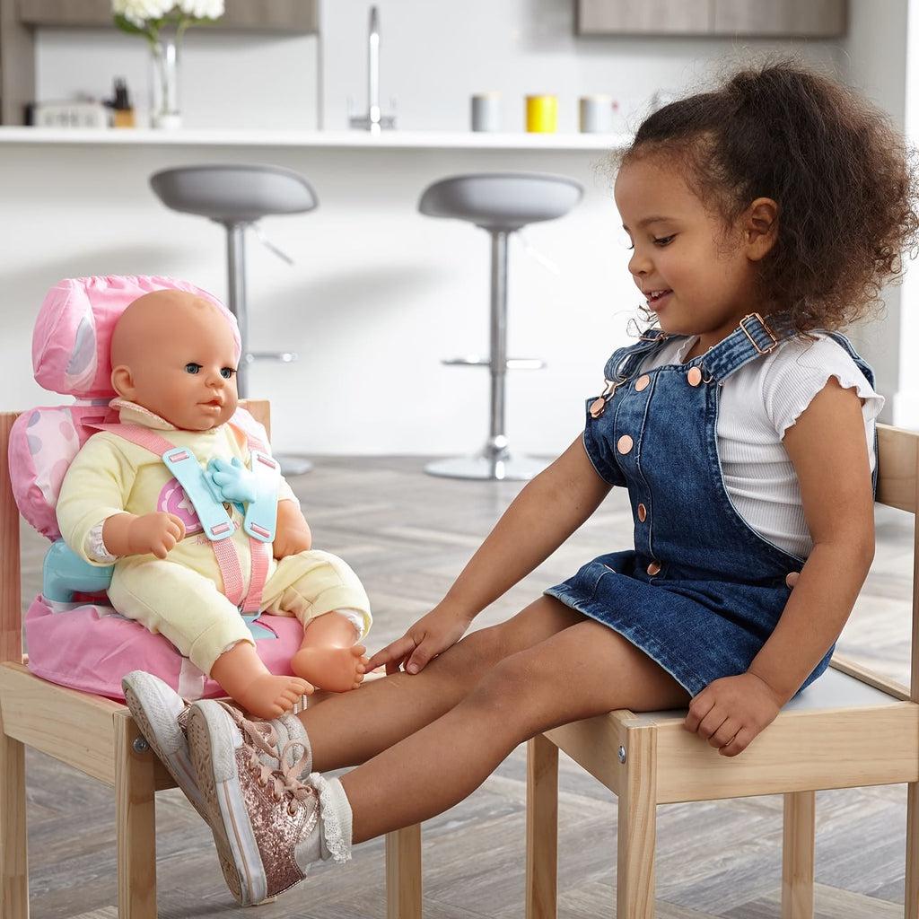 A girl is shown sitting in a char next to another chair where the booster seat is with a baby doll strapped in.