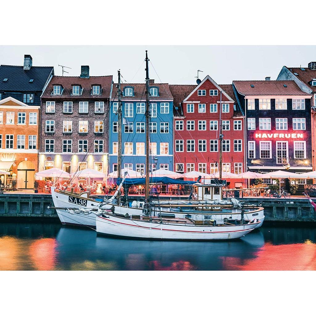 Puzzle image | Colorful waterfront buildings sit behind two boats floating on reflective water | Along the boardwalk between the buildings and water, people dine under umbrellas. | Scene is set at dusk so lights and colors reflect off water.