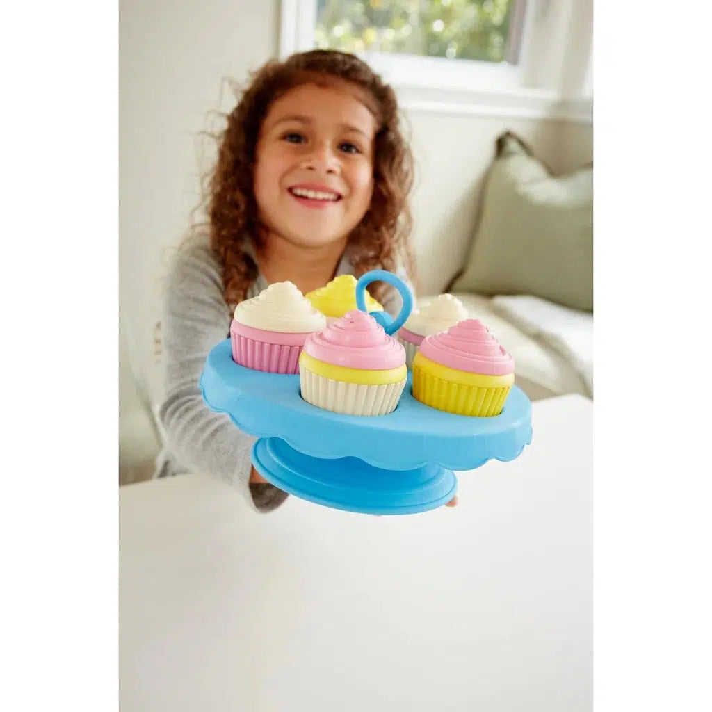 Scene of the same little girl smiling and offering you some cupcakes.