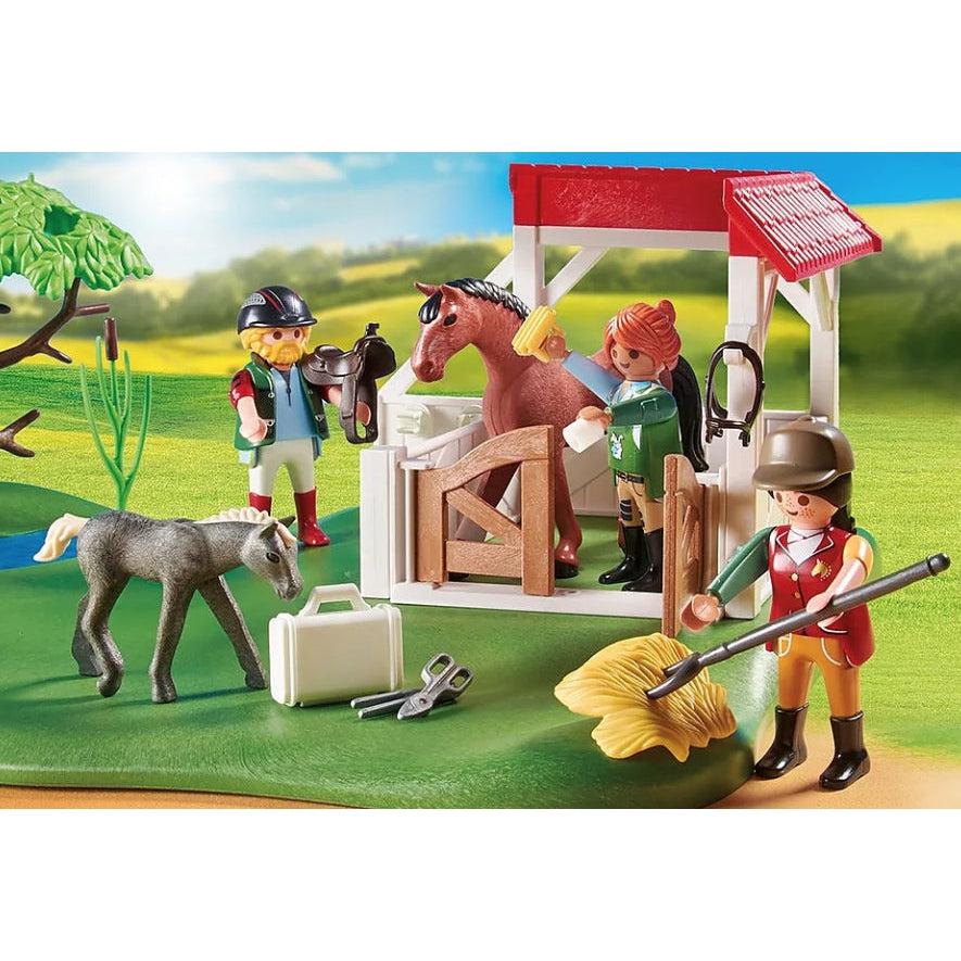 A stabled playmobil horse is being brushed by a playmobile figure while another figures pokes hay with a pitchfork, another figure brings a saddle, and a playmobil foal sniffs at a grooming bag