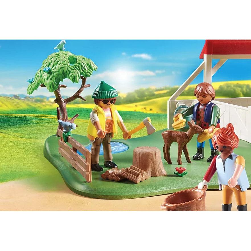 A playmobil figure stands next to a stump and a pile of fake firewood and holds a toy ax, another figure grooms a playmobil fawn and another figure holds a playmobil basket.