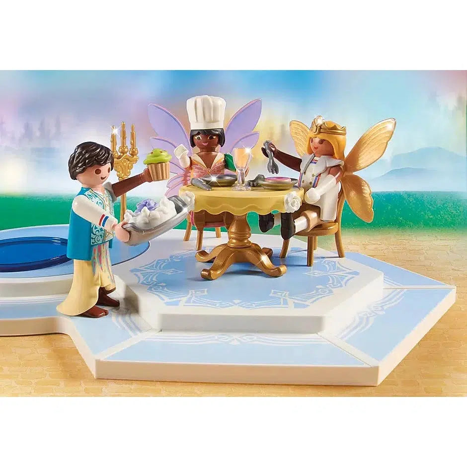 One of the figures brings toy food to two other figures sitting at the table, they both have fairy wings