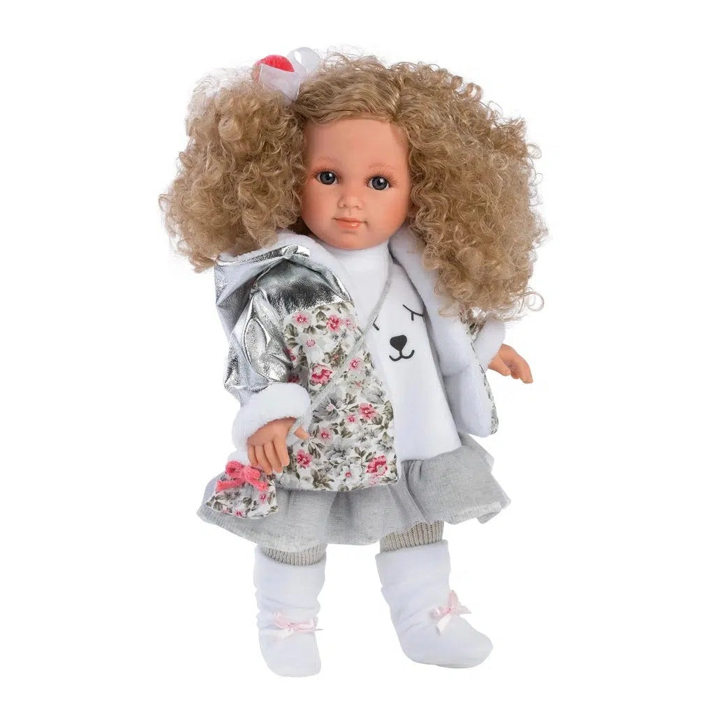 Doll out of packaging | Doll has elbow length curly blonde hair, and blue eyes. Her hair has a white and red bow in it. She is wearing a white shirt with a graphic design resembling a cats face. She has on a jacket that is silver metallic and gray/pink floral with a white fur lining. She has on a gray skirt and leggings, and white socks with pink bows. She has a purse that matches the floral print of her coat.