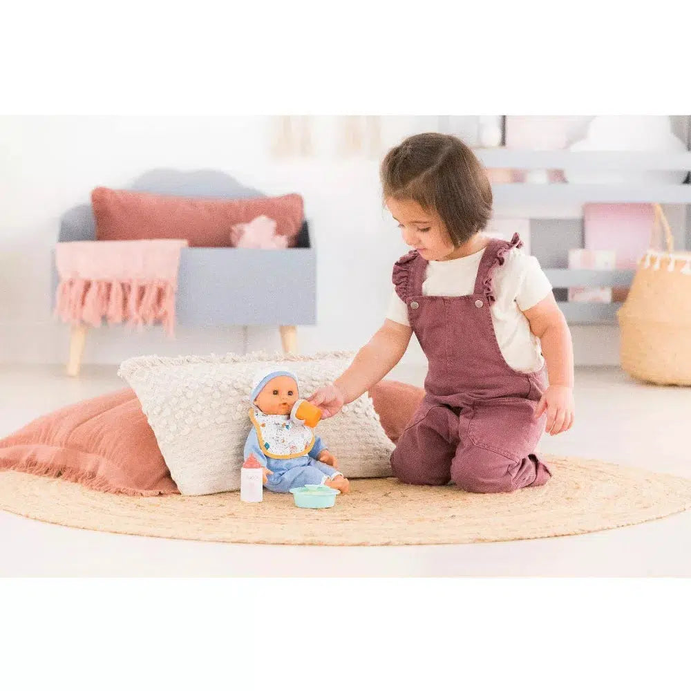 A girl is shown holding the sippy cup up to the mouth of her doll which is wearing the bib and sat next to the bottle and bowl that are also in the set.