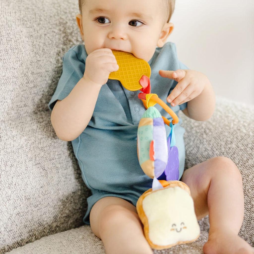 Infant sits holding toy with brown plastic peanut portion in mouth