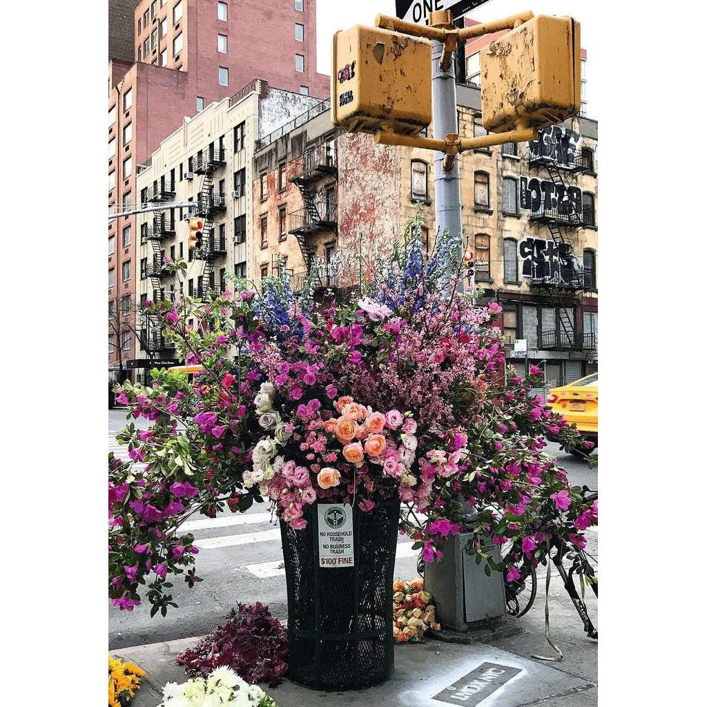 Puzzle image | Expensive flower display sits in a city street trashcan on a New York street corner. Behind the flowers a weathered crossing light and buildings with graffiti.