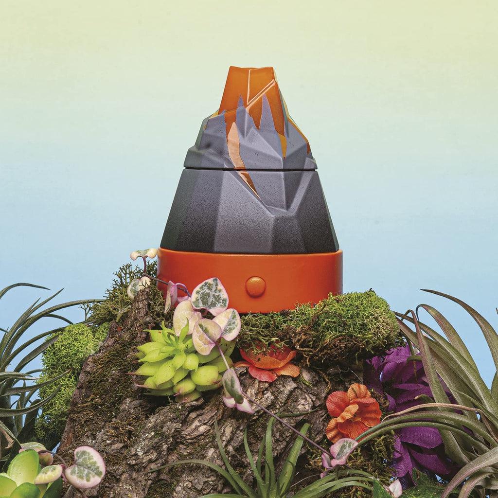 Image of toy fully assembled. | Toy has an orange base with button, upper portion of toy is gray and orange plastic shaped like an erupting volcano. | Toy shown against light blue background surrounded with tropical plants and moss.
