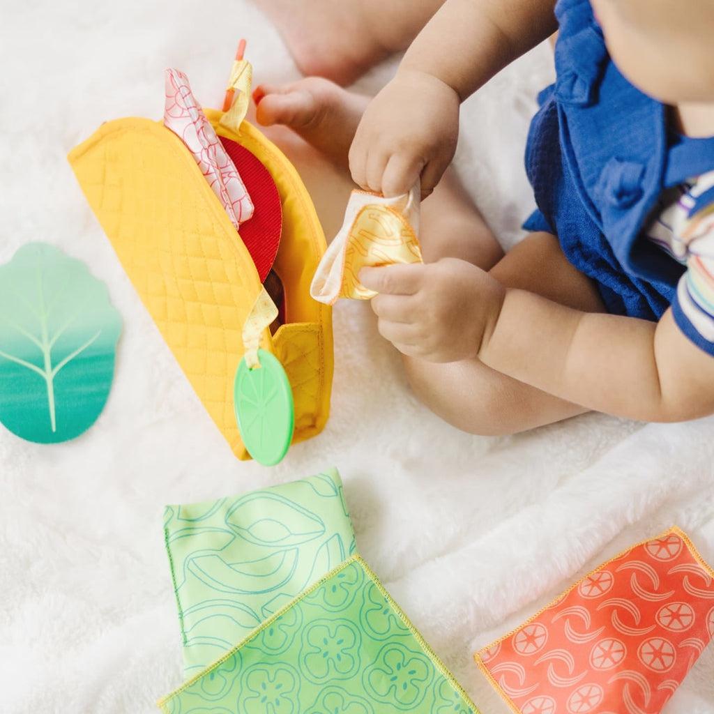 Infant sits on floor playing with toy holding soft fabric piece over foam taco shell
