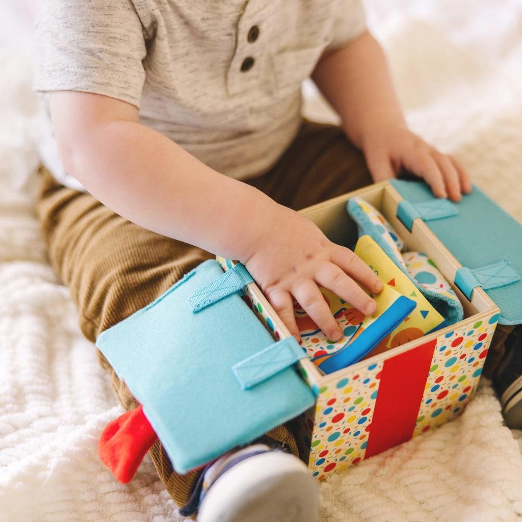 Infant with toy in lap opens blue flaps of wooden git box to reveal contents inside