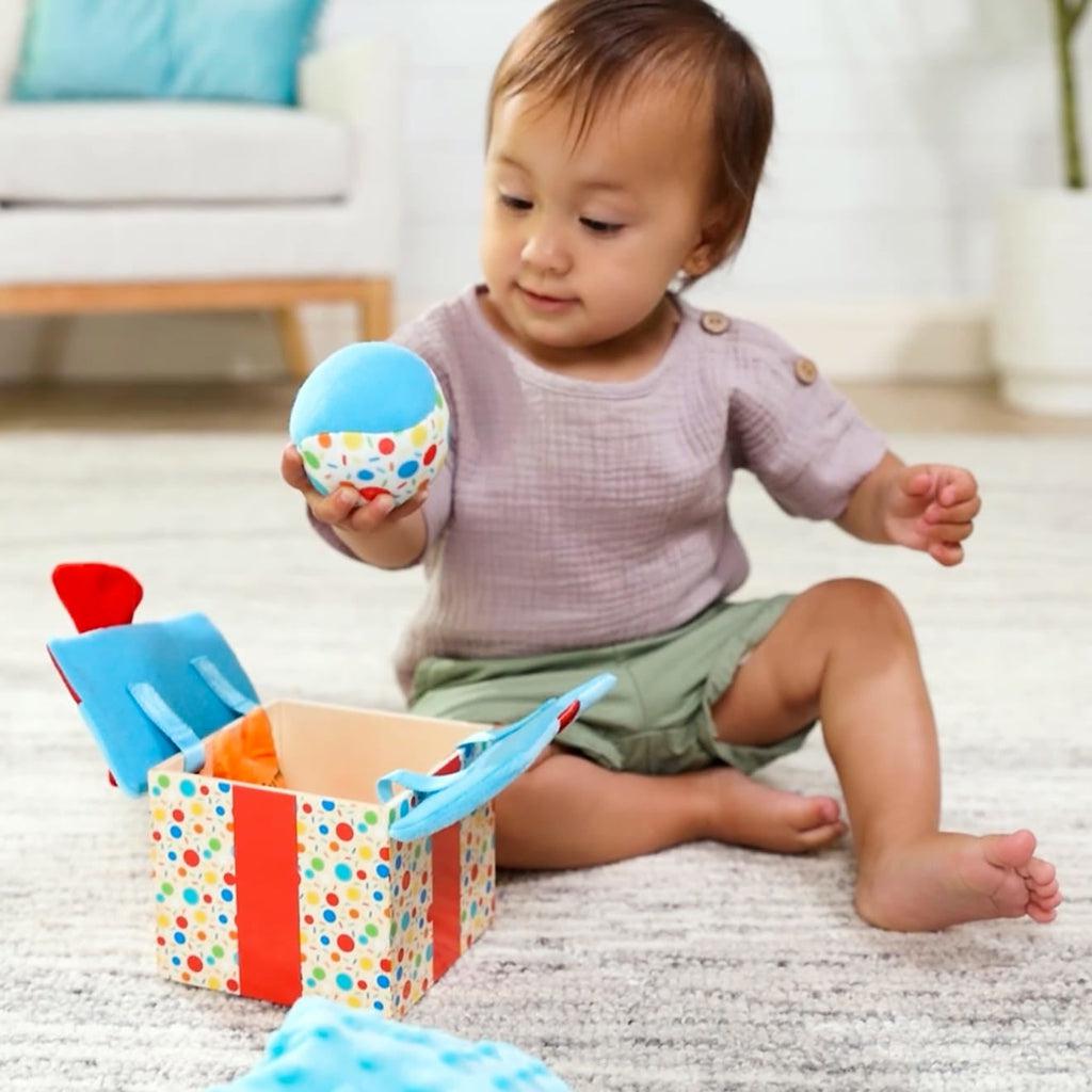 Infant sits on floor with wooden gift box open | Infant is holding the squeezable ball
