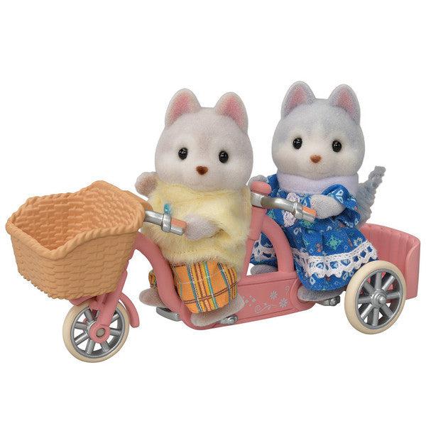 Shows an up close view of the two siblings on the pink tandem bicycle. It has a basket in the front. The sister is wearing a blue dress and the brother is wearing a yellow sweater and orange plaid pants.