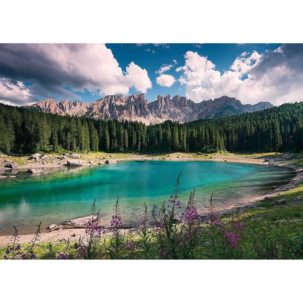 Puzzle image | A turquoise lake sits surrounded by mountain greenery with purple flowers in front and a vast expansion of pine trees behind | Mountain peaks and a blue sky are visible above the tree line.