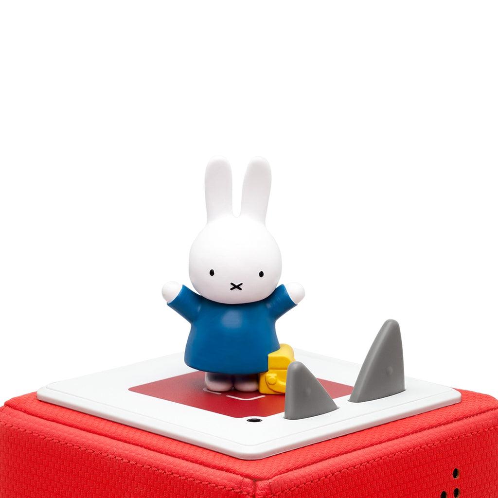 The tonie is shown on a red tonie box. the figure is a white rabbit on two legs holding it's arms up with a navy blue dress/oversized shirt. There is a yellow bag at its feet