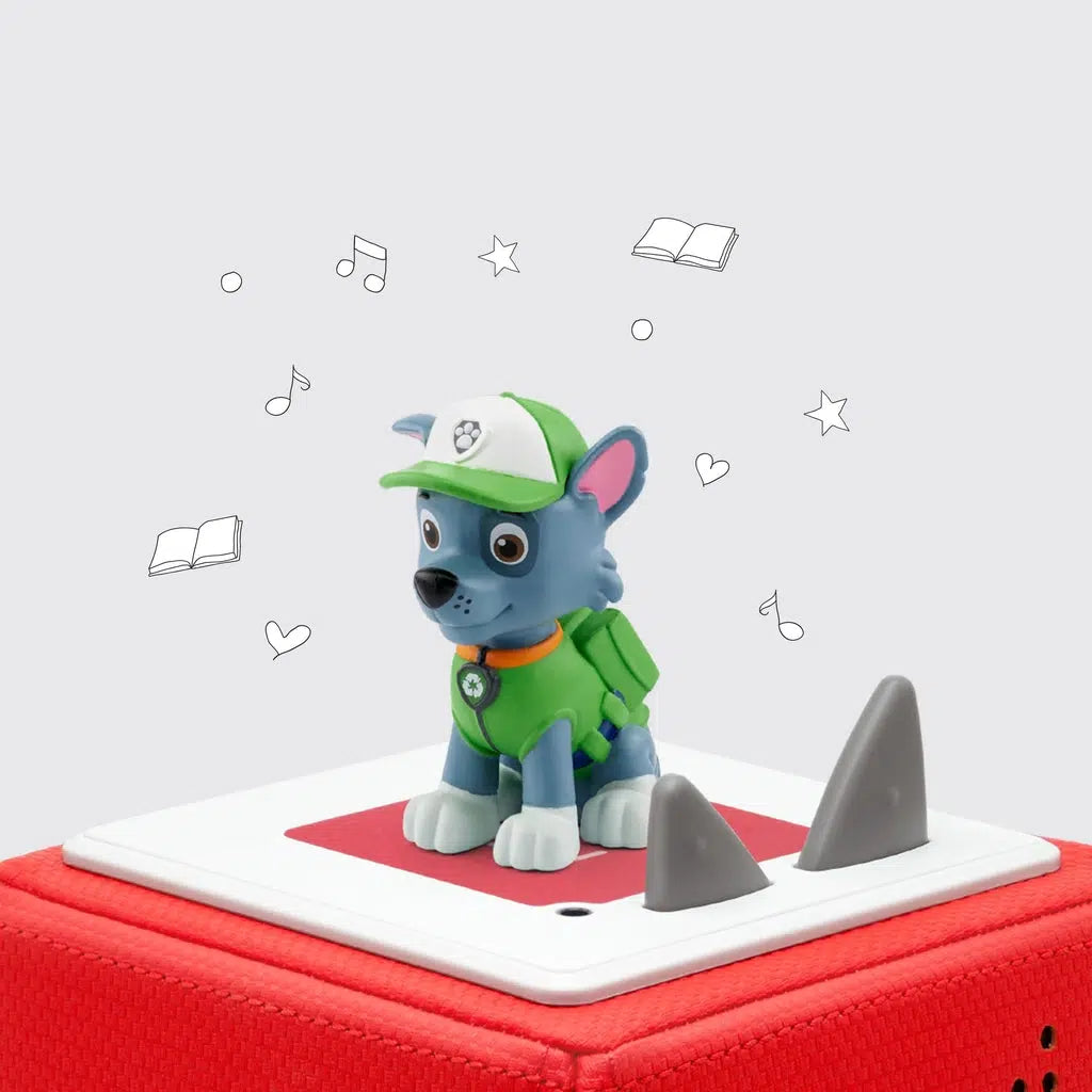 tonie figure on a red toniebox | figure is a grey dog with white paws and a dark spot around one eye, it's wearing a green vest, hat, and backpack, and a recycling symbol on the dog tag.