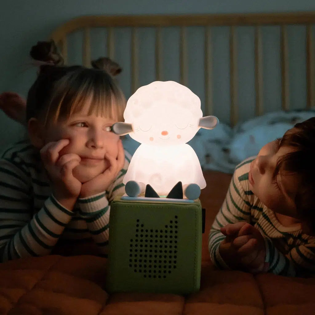 two children smile at the night light tonie that's glowing on top of a green tonie box in a dark room on a bed