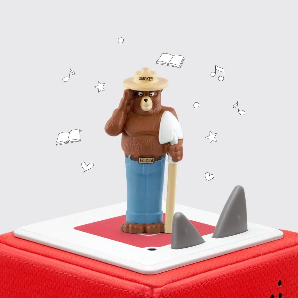 The tonie is smokey bear, a bear wearing blue jeans, a buckle with his name, a wide brimmed hat, and a shovel, standing on two legs