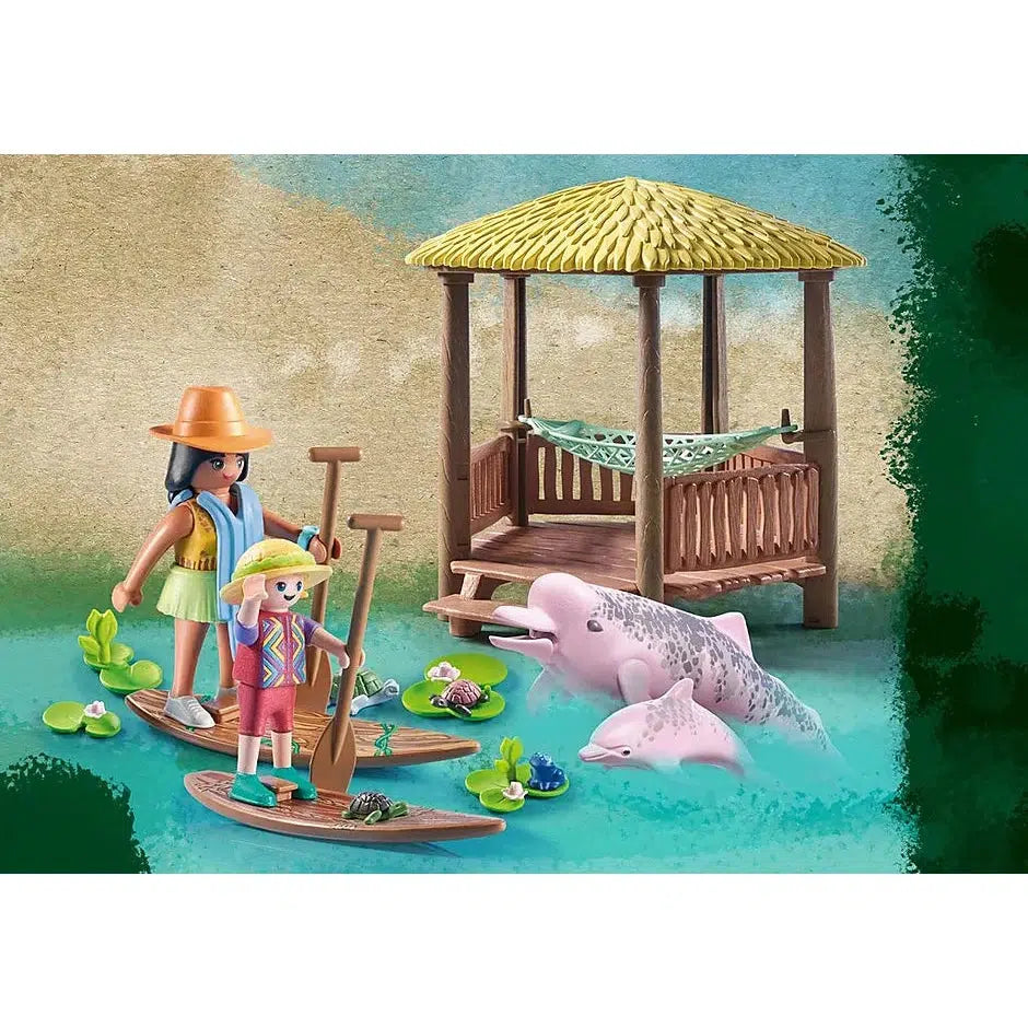 The playset is shown, two figures, one mother and one daughter, stand on paddle boards looking at two pink dolphins, one adult and a baby, floating in a river next to a veranda with a hammock in it